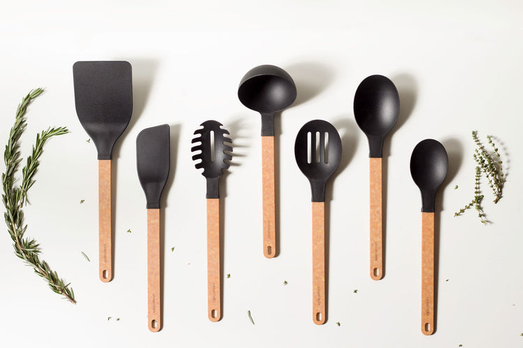 A group shot of various utensils from our lines on a flat counter next to some fresh herbs
