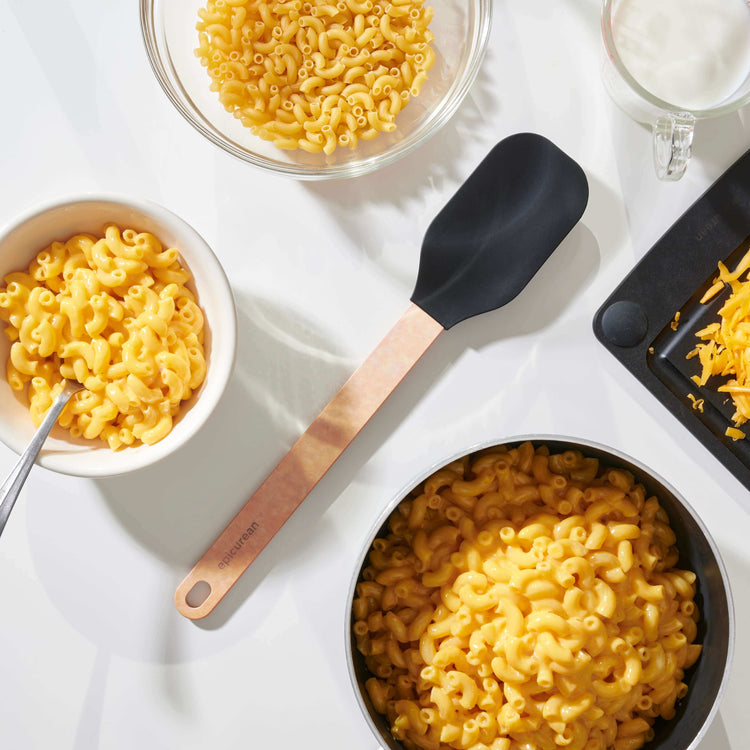 Epicurean Silicone Series Large Spoonula in Natural/Black on a white counter surrounded with bowls of ready to eat macaroni and cheese with all of the ingredients to prepare more
